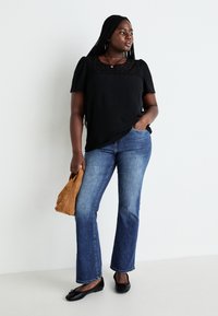 Black top with lace detail, blue flared jeans, black flat shoes, and brown suede bag. Model stands against a white background.
