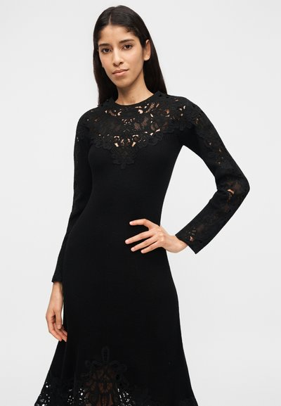 Γυναίκα με μακριά σκούρα μαλλιά, φορώντας μαύρο φόρεμα μακρυμάνικο με περίπλοκες δαντελένιες λεπτομέρειες στο στήθος, τα μανίκια και το τελείωμα, ποζάροντας με το ένα χέρι στη μέση.