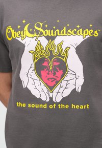 Camiseta gris con un diseño gráfico: manos sosteniendo un corazón con un rostro, llamas y el texto "Obedece Paisajes Sonoros" en amarillo, con acentos en rosa.