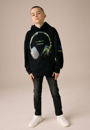 Garçon debout face à nous, portant un sweat à capuche noir avec un motif coloré de casque audio, un jean foncé et des baskets blanches sur un fond uni.