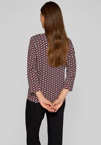 Dreiviertelärmeliges Shirt mit einem geometrischen Muster in Rot, Weiß und Schwarz. Glattes Material mit rundem Ausschnitt und lockerer Passform.