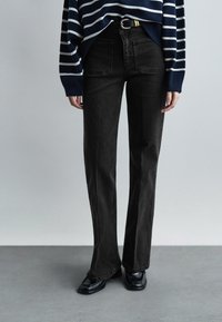 Άτομο που φοράει μαύρο τζιν ψηλόμεσο, μαύρη ζώνη, μπλε σκούρο και λευκή ριγέ μπλούζα με μακριά μανίκια και μαύρα παπούτσια με τετράγωνη μύτη σε γκρι δάπεδο.