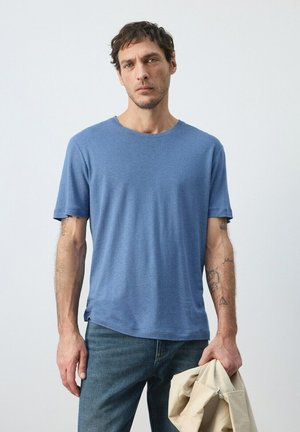 Uomo con capelli corti e scuri, che indossa una maglietta blu e jeans, tiene una giacca beige nella mano destra, in piedi davanti a uno sfondo chiaro e uniforme.