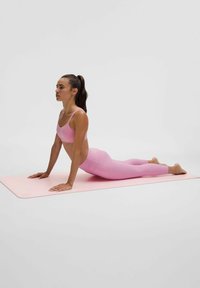 Roosa spordiriietes naine teeb joogaasendit "ülespoole koer" roosal joogamattal neutraalse tausta ees.