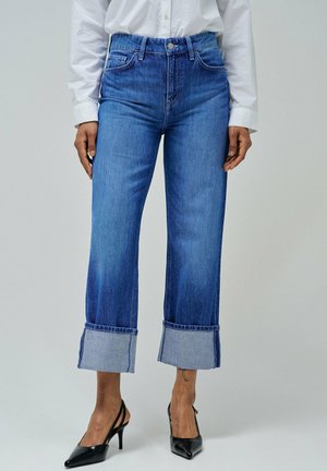 Jeans de mezclilla de cintura alta en azul, con piernas anchas y dobladillo con puño, que cuentan con costuras en contraste. Llevados con tacones negros tipo slingback.