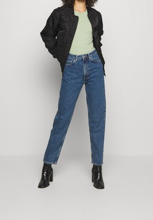 Chaqueta bomber negra, camiseta verde claro, jeans de mezclilla azul de cintura alta y botines negros de tacón. Tejido suave, corte estructurado.