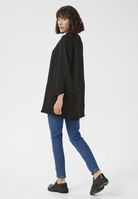 Svart oversized topp med 3/4 ärmar, texturerad tyg, ihop med figurnära blå jeans och svarta platåsandaler, vy från baksidan.