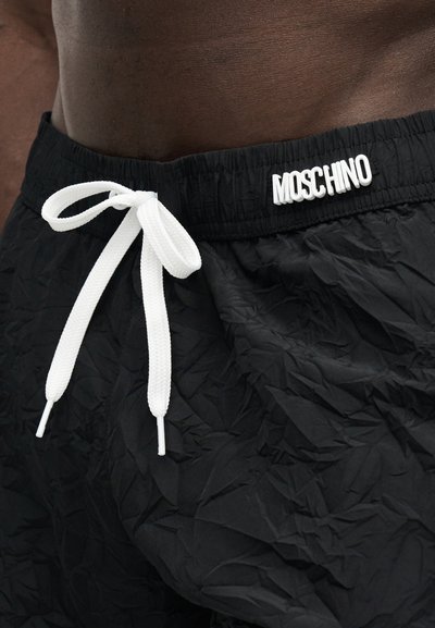 Μαύρα τσαλακωμένα υφασμάτινα σορτς με λευκό κορδόνι δεμένο σε φιόγκο και λογότυπο "MOSCHINO" στη ελαστική μέση, φορεμένα σε σκούρο δέρμα κορμού.