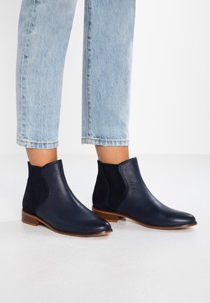 Personne portant des bottines en cuir bleu marine avec des panneaux latéraux élastiques noirs et des semelles en bois, associées à un jean court bleu clair.
