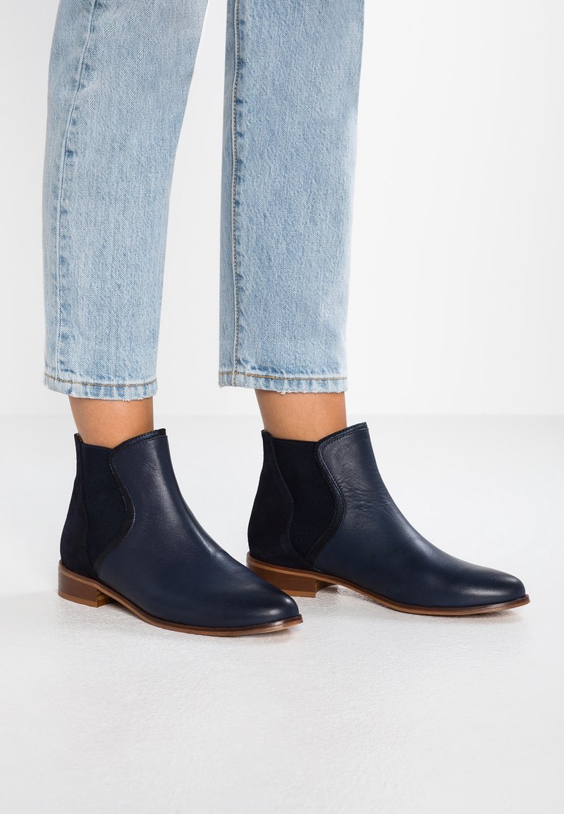 Personne portant des bottines en cuir bleu marine avec des panneaux latéraux élastiques noirs et des semelles en bois, associées à un jean court bleu clair.