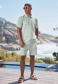 Polo shirt verde chiaro con motivo e pantaloni corti abbinati, realizzati in tessuto testurizzato. Il modello indossa sandali marroni e occhiali da sole, in piedi all'aperto.