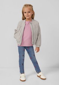 Ragazza giovane con giacca grigia con zip, maglietta rosa, jeans blu con motivi a fiocco e sneakers bianche, in piedi contro uno sfondo semplice.
