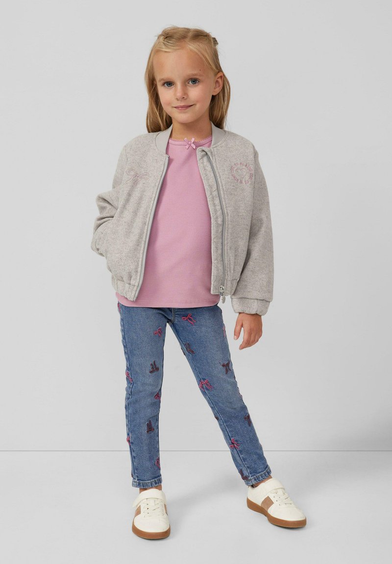 Ragazza giovane con giacca grigia con zip, maglietta rosa, jeans blu con motivi a fiocco e sneakers bianche, in piedi contro uno sfondo semplice.