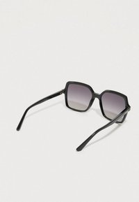 Černé sluneční brýle ve tvaru čtverce s gradientními čočkami, tlustými plastovými obroučkami a štíhlými stranami. Výrazným detailem na straně je značka.