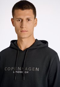 Sudadera negra de mezcla de algodón, con bolsillo tipo canguro, cordones y "COPENHAGEN LINDBEGH" impreso en letras bold en la parte frontal.