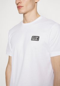 Camiseta de algodón blanca con un parche bordado del logo EA7 en negro en el lado izquierdo del pecho. Manga corta y cuello redondo. Textura suave.