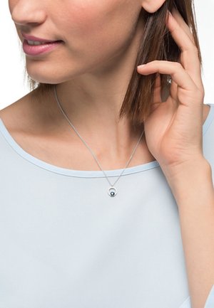 Silberne Halskette mit einem runden Anhänger, der einen kleinen, reflektierenden blauen Stein zeigt. Getragen von einem Model mit einem hellblauen Oberteil.
