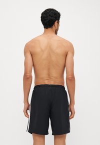 Svarte svømmebukser laget av lett materiale, med elastisk midje og hvite side striper, vist fra bakvinkelen.