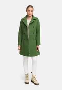 Manteau vert croisé avec un col cranté, featuring des boutons noirs, des poches latérales et une coupe ajustée, porté sur un top beige et un pantalon blanc.
