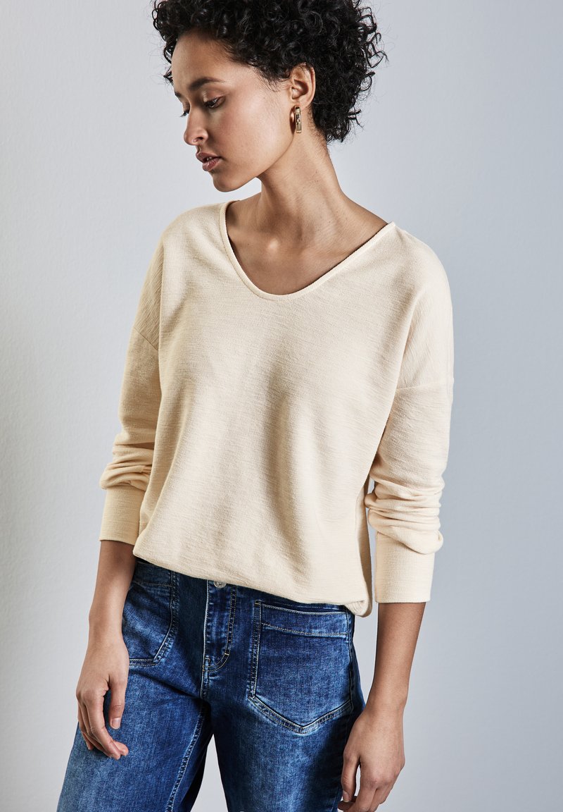 Maglietta beige chiara a maniche lunghe con scollatura ampia, tessuto testurizzato e vestibilità rilassata, abbinata a jeans in denim blu con tasche posteriori.