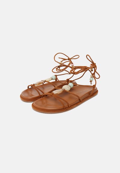 Sandales plates marron avec fines lanières, liens à la cheville et grosses perles décoratives sur les lanières avant sur fond blanc.