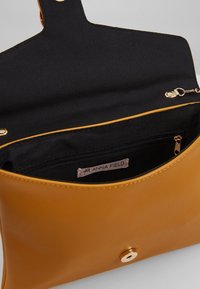 Musztardowa skórzana torba o gładkim wykończeniu, wyściełana czarną tkaniną. Posiada kieszeń na zamek oraz magnetyczne zapięcie na zatrzask.