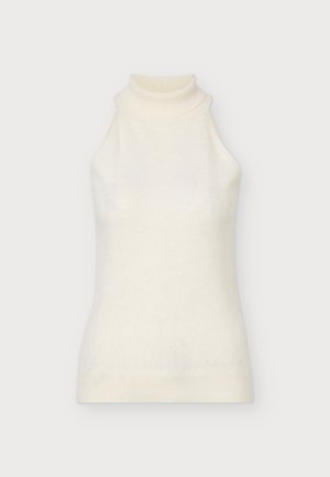 Le modèle se tient face à l’objectif, vêtu d’un haut crème sans manches à col roulé et d’un pantalon à jambes larges, avec des chaussures noires à bout ouvert, sur un fond gris uni.