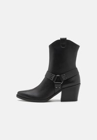 Black leather ankle boot with a pointed toe, stacked heel, and a strap with a metal ring and studs around the midsection.