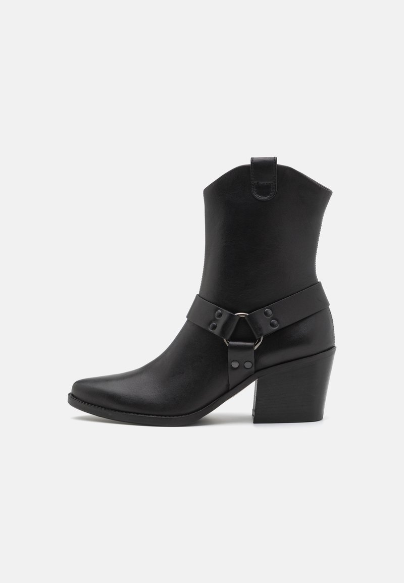 Black leather ankle boot with a pointed toe, stacked heel, and a strap with a metal ring and studs around the midsection.