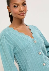 Hellblauer Cardigan mit gerippter Textur, Wellensaum am Ausschnitt und drei dekorativen silbernen Knöpfen. Weiches Material und lange Ärmel.