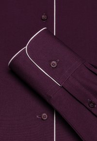 Paarse shirt met witte piping, voorzien van twee knopen op de kraag en manchetten. De stof heeft een gladde, gestructureerde uitstraling.