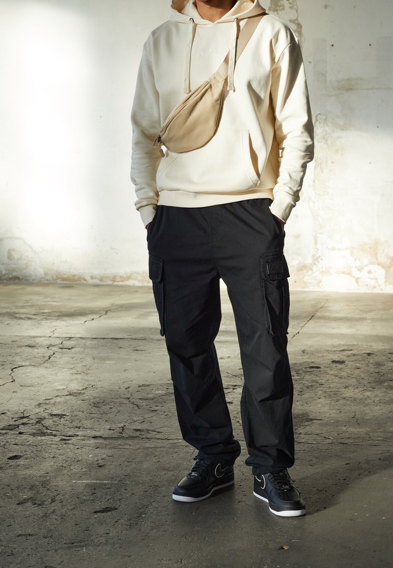 Beige mikina s kapucí a přední kapsou, černé cargo kalhoty s bočními kapsami a černé tenisky. Neformální outfit s kontrastem v barvách a materiálech.