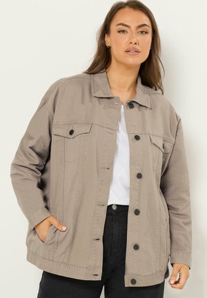 Kvinde iført oversized taupe knappet jakke over hvid skjorte og sorte bukser, stående mod en ensartet lys baggrund.