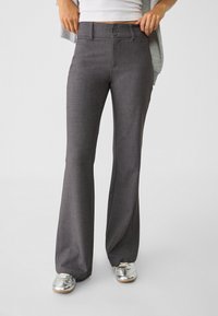 Pantalons évasés gris en tissu texturé, avec une fermeture à deux boutons et des poches avant, associés à des chaussures de sport argentées.