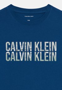 Mėlyna medvilninė marškinėliai su dideliu, tekstūruotu baltu piešiniu priekyje, kuriame kelis kartus ryškiomis raidėmis kartojamas užrašas „CALVIN KLEIN“.