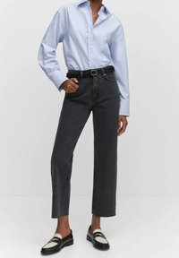 Camisa de manga larga azul claro metida dentro de jeans de tiro alto de color gris oscuro. Cinturón negro con hebilla dorada, mocasines planos en negro y blanco.