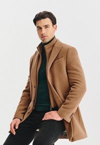 Jasnobrązowy wełniany płaszcz z wysokim kołnierzem, zapinany na jeden guzik, z bocznymi kieszeniami, noszony na ciemnozielonym golfie, w połączeniu z czarnymi spodniami.