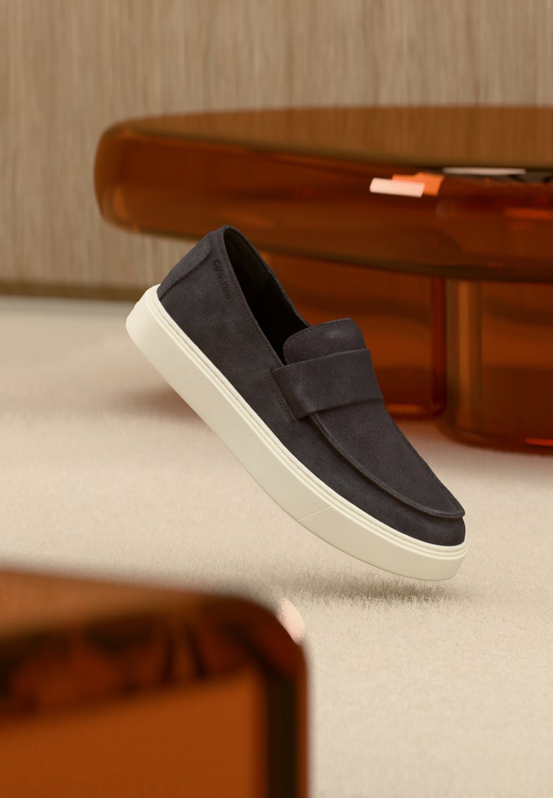 Γκρι σκούρο σουέτ παπούτσι χωρίς κορδόνια με λευκή σόλα, αιωρούμενο πάνω από μπεζ χαλί, με έπιπλα διαφανή σε κεχριμπαρί απόχρωση στο φόντο.