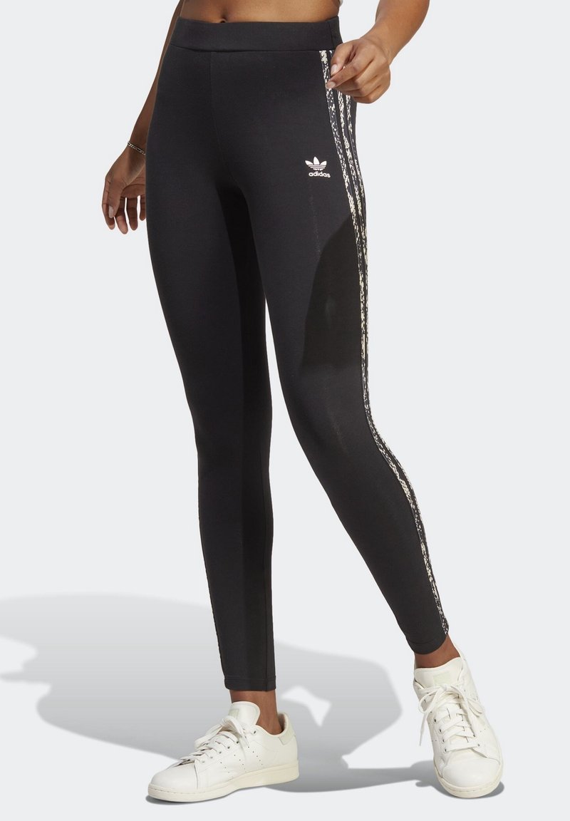 Benen bär svarta Adidas leggings med mönstrade sidostrickor och vita Adidas sneakers, stående på en ljus bakgrund.