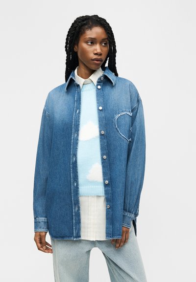 Femme aux longs cheveux tressés portant une chemise en jean surdimensionnée par-dessus un pull bleu clair avec des motifs de nuages blancs et un pantalon de couleur claire.