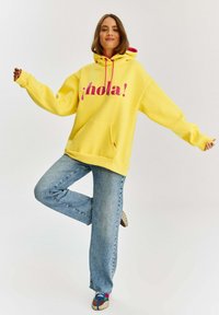 Žltá mikina s kapucňou a nápisom "¡hola!" v ružovom písmene, dlhé rukávy, veľká predná vrecko, skombinovaná s svetlo modrými rovnými džínsy a viacfarebnými topánkami.