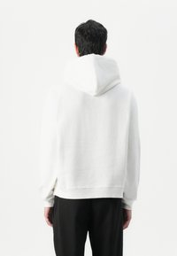 Sudadera blanca de algodón con un corte relajado, que presenta una gran capucha y puños y dobladillo acanalados. La tela tiene un acabado suave y texturizado.