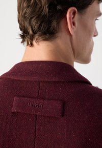 Palton dintr-un amestec de lână de culoare burgundă, cu guler cu vârfuri, având o finisare texturată și o etichetă cusută cu inscripția "HUGO" pe spatele gâtului.