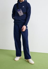 Mörkblå hoodie och byxor i mjukt material, med vita sidoflikar, tillsammans med vita skor och en liten grå axelväska.