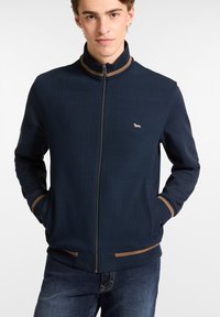 Mornarčka zip-up jakna s teksturiranim prednjim dijelom, glatkim rukavima i svijetlosmešnim naglasnim trakama na ovratniku i manžetama; sadrži mali logotip na prsima.