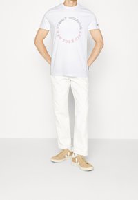 Camiseta blanca de algodón con un estampado gráfico circular en rojo y azul que dice "TOMMY HILFIGER NEW YORK CITY", combinada con zapatillas beige y pantalones blancos.