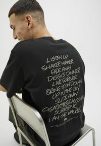 Black cotton t-shirt featuring large, handwritten-style text printed in light gray on the back, with various song titles listed vertically.