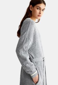 Cardigan grigio lavorato a maglia con cintura a cravatta, caratterizzato da polsini a coste e sottili motivi testurizzati sul tessuto. Materiale morbido e accogliente.