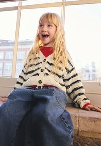 Fille aux longs cheveux blonds portant un pull rayé et un jean, assise près d'une grande fenêtre, regardant vers le haut et souriant.