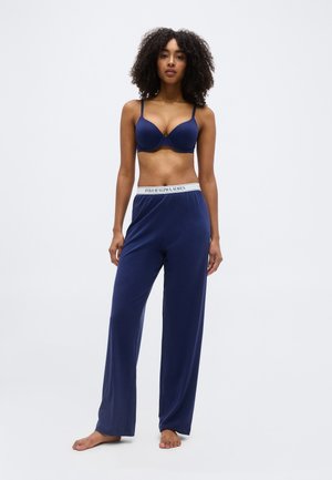 Femme aux cheveux bouclés portant un soutien-gorge bleu marine et un pantalon large assorti avec une ceinture "Polo Ralph Lauren", debout pieds nus sur fond blanc.
